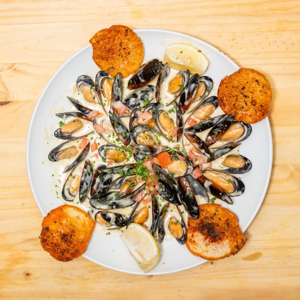 Mussels in a creamy sauce with chopped tomatoes, parsley, lemon, and toasted bread on a white plate.