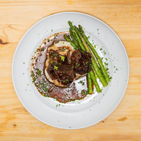 Braised short ribs on mashed potatoes with asparagus and gravy, garnished with parsley.