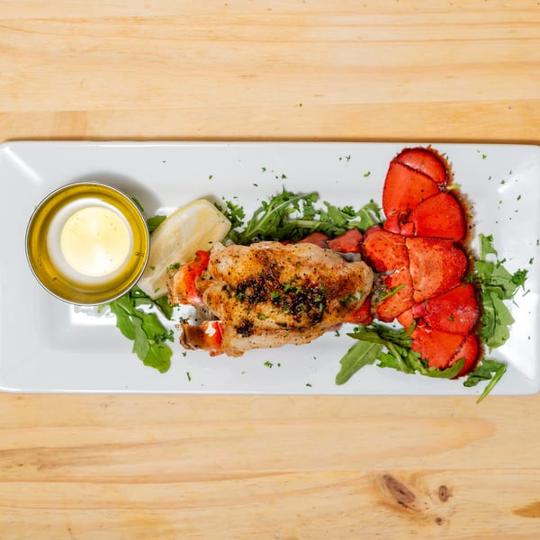 Grilled lobster tail with lemon, butter sauce, and arugula on a white rectangular plate.