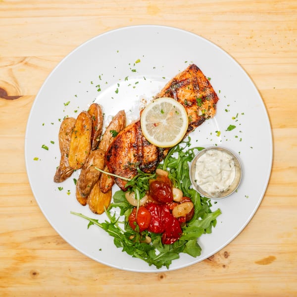 Grilled salmon with roasted potatoes, cherry tomatoes, arugula, and a side of tartar sauce.