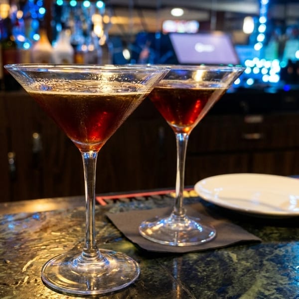 Two martini glasses with dark red cocktails on a marble bar top with blurred bottles in the background.