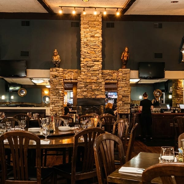 Warmly lit restaurant dining room with dark wood tables, chairs, and a central stone fireplace.