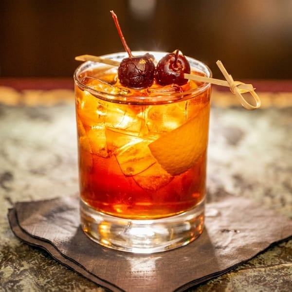 A vibrant amber cocktail with ice, orange peel, and two cherries on a skewer, on a dark napkin.